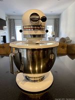 Front view of white mixer atop countertop showing KitchenAid logo, chrome lock knob and attached stainless steel bowl