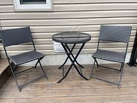 Front view of outdoor bistro set on wooden deck against siding: round glass top table and two folding chairs with woven seats and backs