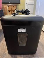 Front view of black paper shredder showing power cord and viewing window for shredded paper bin.