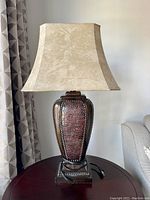 Front view showing the lamp's beige fabric shade and textured faux leather base with nailhead trim on a dark wood or metal pedestal base.
