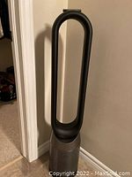 Dyson bladeless tower fan upright against wall, showing silver perforated base and black loop amplifier