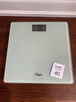 Top view of the Ozeri digital bathroom scale on a wood floor, showing the glass platform and LCD display reading '0.0'.