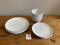Set of white ceramic plates and bowls arranged on a wooden surface; includes 5 small plates stacked, 4 large plates stacked, and 4 small bowls stacked.