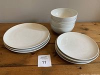 Shows 3 large plates stacked on the left, 4 small plates stacked on the right front, and 4 mid-size bowls stacked on the right back, placed on a wooden surface against a plain wall.