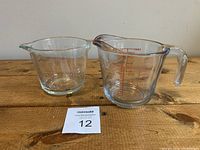 Two glass measuring cups on a wooden surface, one with clear red measurement markings, the other with faded markings but legible.
