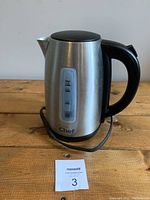 Front view of stainless steel electric kettle with water level indicator and black plastic handle, sitting on wooden surface.