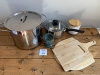 Full lot view with all items on wooden surface: 2 chopping boards, large stainless steel pot with lid, smaller pot with glass lid, white ceramic container with wooden lid, green marble pestle