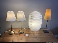 Full view of all four lamps on a wooden surface including two matching brass base lamps with pleated shades, a large white paper lantern style lamp, and one yellow lamp with a slim base.