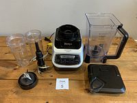Front view of Ninja blender base, large pitcher, two smaller cups with lids, an additional blade, and large lid on wooden surface