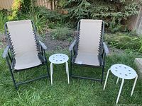 Two outdoor folding chairs with beige fabric seats and black metal frames, alongside two small white round stools or side tables on grass.