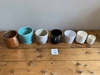 Row of seven assorted planters of varying sizes and designs placed on wooden surface against a plain background. Shows copper, turquoise, white, black, and textured white planters.