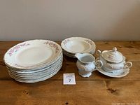 Photo showing 8 dinner plates stacked, 5 bowls stacked, 1 saucer on top and 1 teapot with lid, 1 creamer on wooden table.