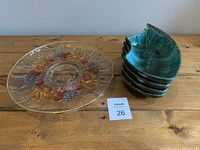 Photo of one large clear glass serving platter placed beside four stacked blue/green glass serving dishes on a wooden table.