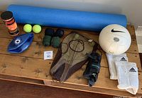 Overview of all sports equipment items arranged on wooden surface including foam roller, volleyball, knee pads, wrist wraps, wrist brace, tennis balls, and paddleball set.