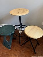 Three stools shown side by side, including adjustable IKEA stool with round light wood top and black metal base, green folding stool with carrying hole, and black folding stool with light wood seat.