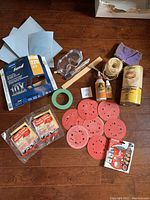 Photo shows multiple woodworking and finishing supplies on wooden floor including sandpaper sheets, sanding discs, safety goggles, green masking tape, twine, glue bottles, paintbrush, and cans of finishing products.