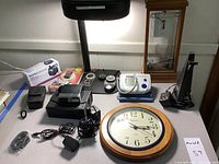 Desk clock with wood frame, battery wall clock, blood pressure cuff, desk lamp, compass collection, Vivitar flash, and cables