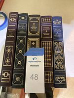 Spines of five hardcover books: Treasure Island, Fathers and Sons, Don Quixote de la Mancha, Madame Bovary, Pere Goriot, all Franklin Library editions with gold detailing on dark covers.