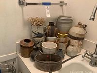 Overview of twelve handmade clay and ceramic pots arranged on a counter near a sink, showing variety in size, shape, and color.