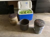 Large blue and white Igloo cooler filled with tennis balls among three storage buckets with lids, one wooden, one metal, and one gray metal with a camo padded lid.