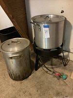 Full view of propane burner stand holding large aluminum fryer pot with lid and smaller fryer pot beside it on floor