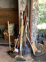 Yard tools leaning against a wall including rakes, brooms, and shovels in garage