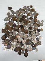 Photo shows a large assortment of British coins spread out on a white surface. Coins range in color from silver to copper tones, with varied designs and conditions visible.