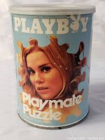 Photo of the original tin container of the 1970 Playboy Playmate Puzzle showing the face of the playmate and puzzle piece design on the lid.