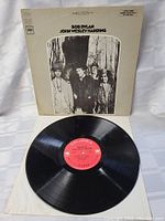 Black vinyl record with red Columbia label alongside album cover showing black and white photo of Bob Dylan and band members, on white background.