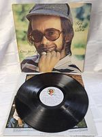 Photo showing the vinyl LP record partially out of the inner sleeve with the album cover behind it, displaying Elton John's face and the album title Rock of the Westies.