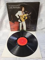 Vinyl LP record outside its cover, showing the red label of the record and the front of the album cover with Paul Simon performing live.