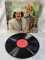 Vinyl LP record and cover of Simon And Garfunkel Greatest Hits shown with cover standing behind record on a white cloth.