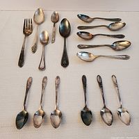 16 pieces of silverplate silverware laid out on a white textured background, including forks, large spoons, and multiple teaspoons with various ornate patterns.