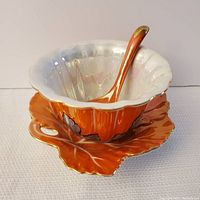 Side view of orange and opalescent hand painted ceramic bowl with matching spoon resting inside on a leaf-shaped dish.