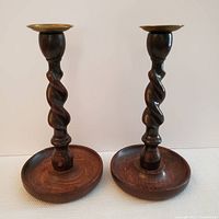 Pair of dark stained wooden Jacobean style candlesticks with barley twist stems and brass cups on top, shown together from a side angle.