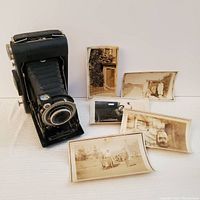 Kodak Vigilant Six 20 bellows folding camera with six vintage black and white photographs beside it on white fabric surface.