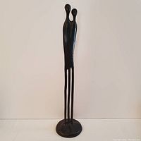 Front view of the dark bronze sculpture depicting two elongated human figures connected, standing on a round base.