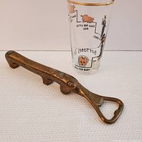 Photo showing the brass dachshund bottle opener and vintage shot glass with gold rim and comical imagery