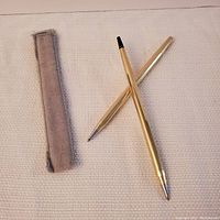 Pair of vintage gold-plated Cross pens, one with the top missing, placed on a white textured surface with a grey fabric sheath beside them.