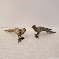 Front view of two metal pheasant figurines with textured feathers and long tails on a light fabric background.