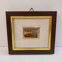 Frontal view of framed small cityscape artwork featuring a canal, bridge and buildings, mounted on fabric backing in wood frame with gold trim.