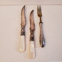 Photo showing three pieces of flatware: two knives with mother-of-pearl handles and one fork with silver handle and brass-colored tines.