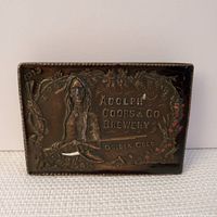 Front view of rectangular bronze finish belt buckle showing embossed Native American figure seated with a bow and surrounding floral motifs, text 'Adolph Coors & Co Brewery Golden Colo.'