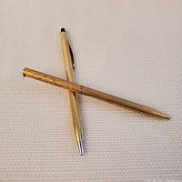 Two gold coloured pens crossed on a light fabric background. The Cross pen shows its textured barrel and gold plated finish. The other pen is smoother and missing its clip.