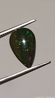 Close-up image of the dark pear-shaped cabochon held by tweezers showing scattered colorful flashes under light.