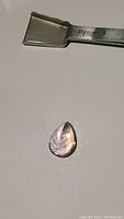 Pear cut ametrine gemstone placed next to metallic object for scale, displaying purple and golden flashes with high luster.