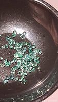 Close-up of numerous small round emerald gemstones, diamond-cut and displaying bright green color, shown in a dark metallic dish.