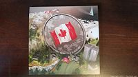 Top view of the coin within a scenic fold-out card showing the Canadian flag and landscape imagery.