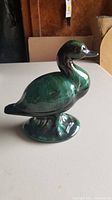 Single ceramic duck figurine in green and black glaze with glossy finish, shown from side angle on white tabletop with neutral background.