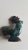 Close-up photo of small ceramic rooster figurine with blue-green glossy glaze on white background.